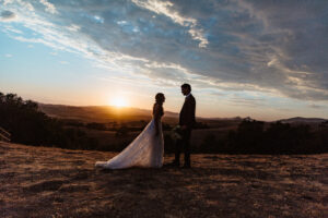 A bride and groom looking into each others eyes on a country side hill. The groom is in a black suit, while the bride is in a white dress. the groom is holding a bouquet of flowers. There are rolling hills in the background while with a sunset cresting over a mountain range far off in the distance.