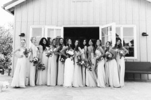 A black and white image of the bride and her brides maid's standing in front of a tall board n bat building with it's French doors open.