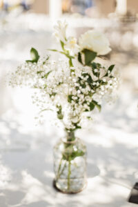 A white rose and small little white flowers in a glass vase resting on a table with a white table cloth.