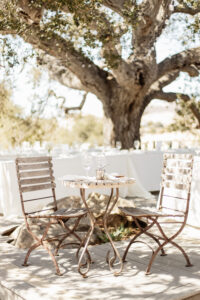 A round table with two old wood/metal chairs on each side with a single candle and two champagne glasses and silverware. In the bright background are rows of tables with white clothes and glassware settings under a large oak tree even further back.