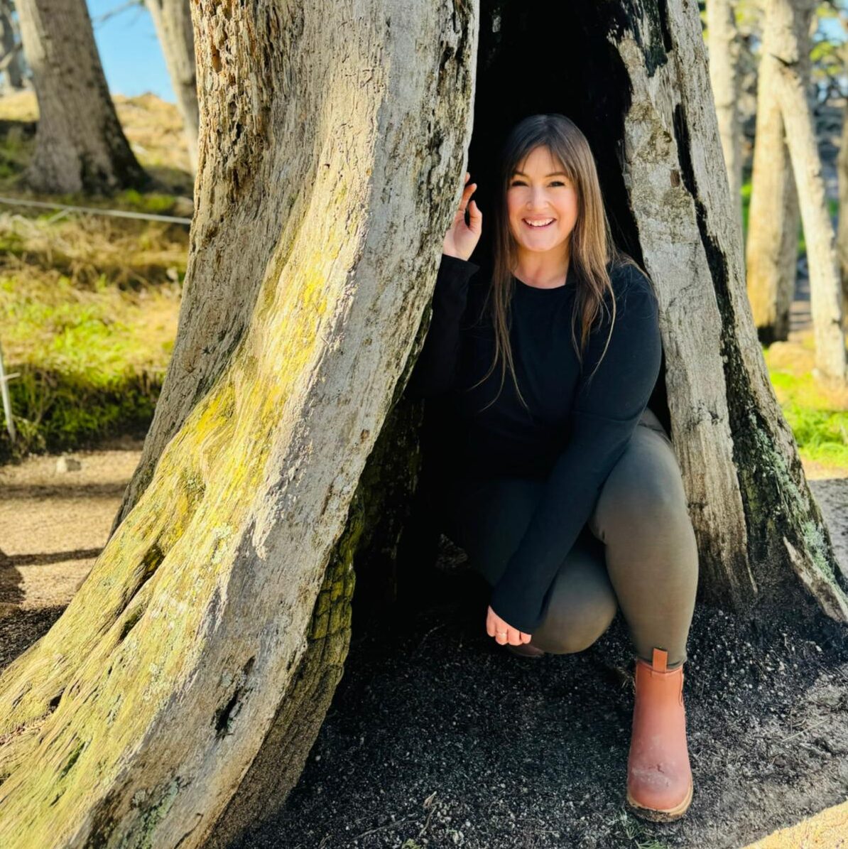A self portrait of Lindsey kneeling down in a hollowed out tree vertical tree trunk. Lindsey had long brown hair with a great smile, she is wearing a long sleeve black shirt, grey jeans, and brown boots.