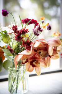 A very colorful flower arrangement with yellow, red, and purple flowers resting in a glass mason jar on a window seal with light shinning through.
