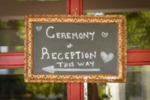 A chalk board with a gold floral metal frame hanging from a red framed glass window. The sign reads; "ceremony and reception this way" with an arrow pointing to the left and hearts scattered around the text.