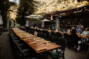An outside dinning seating arrangement of two rows of tables each including; green chairs, yellow napkins, and clear glasses. A group of people are enjoying themselves towards the right side of the image, while workers are spread throughout. It is a wooded environment in the shade of big trees.