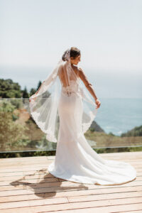 A bride in a white dress standing with her arms out holding her veil. She is standing on a wooden deck that has a glass hand rail over looking a green belt and the ocean.