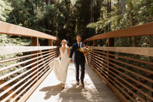 A bride in a white dress and groom dark blue tux holding a bouquet of flowers walking across a wooden bridge over a small creek in a costal forest.