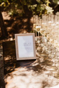 A wooden framed sign on a table with glasses that reads; "Sip Sip Hooray, Ceremony at 3:30"