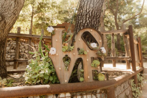 A 3d wooden sign placed in a planter box that says love, but the letters are stacked with the lo over the ve. There are succulents in and around the sign, and there is a big oak tree trunk holding it up. The planter has a metal pipe boarder with chick wire filling in the space. in the background there are trees and a stone wall.