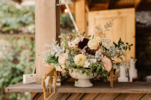 A light pink, red, soft yellow, and white flower arrangement in a white ceramic urn vase resting on a wooden shelf with disposable cameras laying around it.