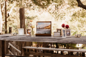 A wooden table with a items on top including: disposable camera, old childhood images in frames, flowers in vases, and a book titled" Big Sur Reveled" resting on a stand. There is a strand of lights hanging from the trees in the background.