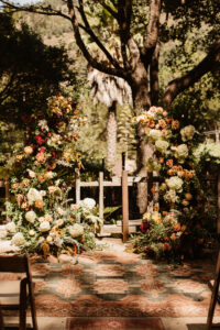 An alter with orange and white flowers on both sides of a rustic rug. Trees and shrubs are shadowing the backdrop.