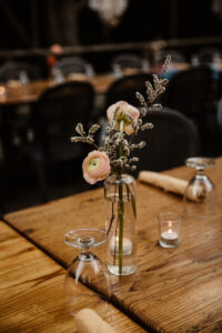 Two pink roses along with two small white flowers in a clear glass vase next to two drinking glasses and a lite candle on a wooden table.