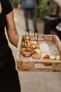 A server holding a tray with samples of food that are rolled up and have twigs sticking out of them like toothpicks.