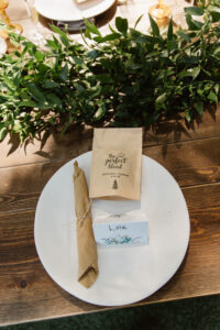 A white plate setting with a bag of coffee, a silverware set wrapped in a brown napkin, and a name tag that reads Lola. The plate is resting on top of a wooden table with a citrus branch and leaves above it.