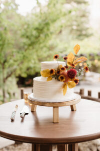 A white wedding cake with yellow and red colored flowers and autumn leaves. The cake is sitting on a three leg stand which is on an old oak table. Green foliage is blurred out in the background.