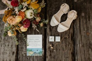 A tan pair of women's dress shoes next to a bouquet of warm colored flowers above a necklace, rings, couplings, and a save the date card.