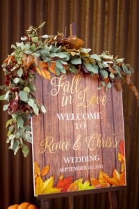 A sign sitting on an easel with eucalyptus leaves hanging on the top of it. The sign reads; Fall in Love, welcome to Rene & Chris's wedding. The easel is standing in front of an rusted metal wall.