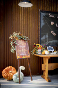 A sign sitting on an easel with eucalyptus leaves hanging on the top of it. The sign reads; Fall in Love, welcome to Rene & Chris's wedding. There is a orange pumpkin, a green pumpkin, and a white squash resting on the concrete ground to the left of the easel. On the right side of the easel is a table with other small pumpkin, and yellow/orange flower arrangements. There is a rusted metal wall in the background with a rustic spot light hanging over head.