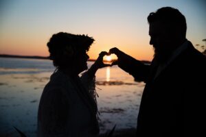 A bride and groom's dark silhouette with each of them holding their hands together in a heart shape with the setting sun in the background. They are over looking a bay of water and the light is reflecting of its surface.