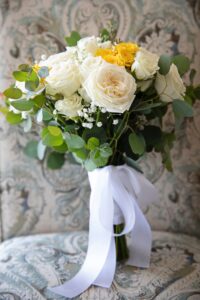 A bouquet of large white roses with medium sized yellow roses mixed in that is held together with a white silk ribbon and is resting on a venetian style chair.
