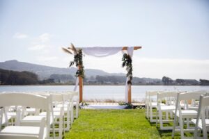 A row of wooden chairs resting on a clean cut grass with a pathway leading to a wooden wedding alter that has tree leaves and feather like plants on its sides and a draping white cloth on top and around it. In the bright daylight background is a large bay of water with tree and hills in the distance.
