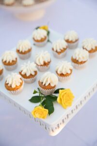 A white box like serving tray holding 13 mini carrot cupcakes with white frosting, and three small yellow roses in two opposite corners.